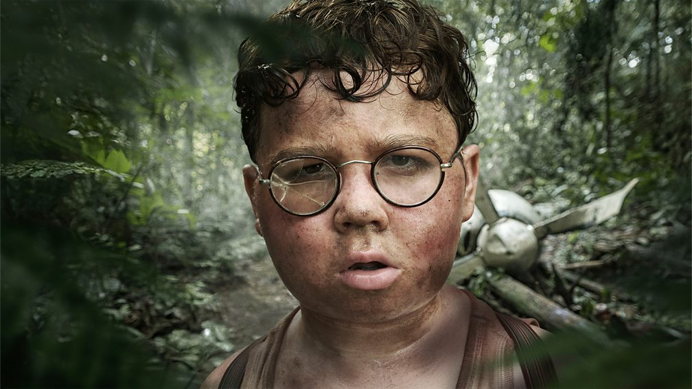 Promotional image for Lord of the Flies showing one of the characters, 'Piggy,' in a jungle staring at the camera. He's dirty and his glasses are broken. There is a crashed plane in the background.