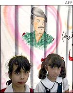 Iraqi girls sit under a drawing of President Saddam Hussein