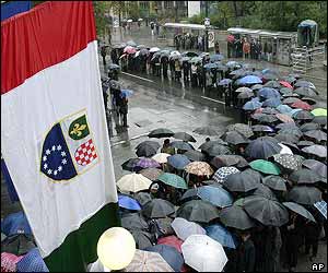 Flags hang in the foreground as people wait under umbrellas to pay their respects to former President Alija Izetbegovic 