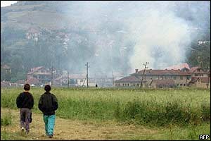 Two young Kosovars walk in a field as several houses burn in Mitrovica