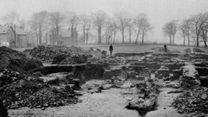 Black and white view of excavations of Roman fort at Mumrills. A line of trees and two large houses can be seen in the background.