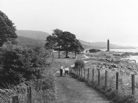 Black and white view of a couple with a pram and dog walking along a winding, coastal footpath. In the background are trees and the tall, cylindrical Battle of Largs Monument.