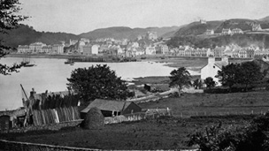 Black and white view of Oban, looking across farmland to the bay, harbour and town.