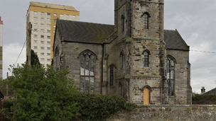 Church with central square tower standing with an open field in the foreground and a high-rise block of flats behind.
