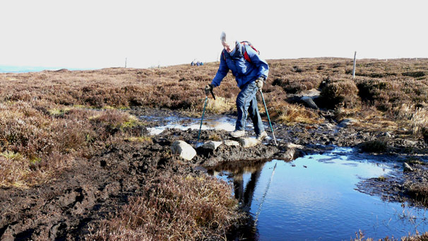 Walker using trekking poles to use stepping stones across mud