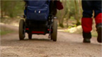 Wheelchair and a walker's feet heading down a forest path.