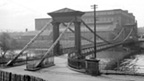 Black and white view of St Andrews Suspension Bridge, a pedestian bridge over the River Clyde. In the background stands a large, industrial building.