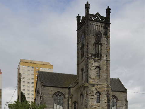 Church with central square tower standing with an open field in the foreground and a high-rise block of flats behind.