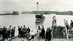 Black and white view of a boat being launched from a slipway into a harbour. A group of men and boys watch from the shore.
