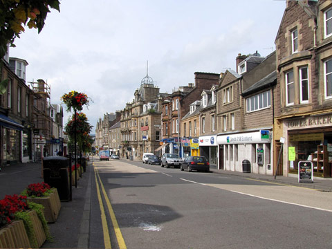 Colour view of Crieff High Street. The street is largely empty, save for a few parked cars and a van driving off into the distance. To the left of frame, the street is lined with ground level and raised planters containing red flowers and foliage.