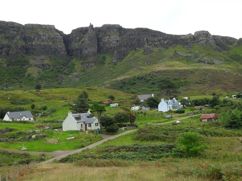 Colour view of a natural rock amphitheatre in which lies a collection of one and two-storey houses along a single-track road.