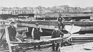 A group of men in Victorian dress stand among boats and carpentry equipment on the shoreline with a row of cottages behind.