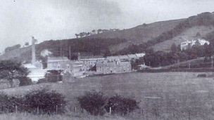 Black and white view of Atheenic Mill buildings and chimney stack, set on a largely grassy slope. There are two short terraces of houses by the mill buildings and a large detached house higher uphill.