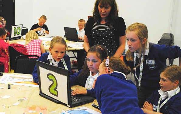girls poring over a laptop in a classroom, watched by an adult