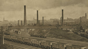 Black and white view of Coatbridge Tinplate Works, dominated by a number of tall, industrial chimney stacks. There is a railway siding in the foreground and the church spires of central Coatbridge can be seen beyond.