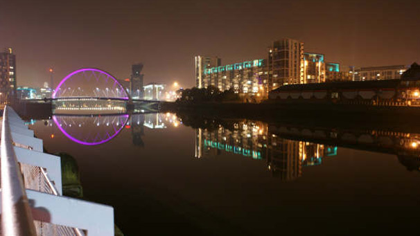 A brightly lit bridge and buildings reflected in the waters of the clyde.