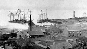 Black and white view over rooftops and industrial chimneys to sailing boats in harbour.