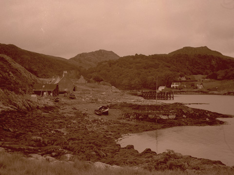Black and white view across bay to a wooden pier with scattered cottages and hills behind. Two boats lie pulled ashore in the foreground..