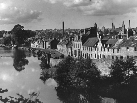 Black and white view of wide flat river with a town to the right of frame. A line of stone, terraced housing lines the street overlooking the river. Several tall industrial chimneys can be seen behind.