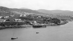 Black and white view across bay to a wooden pier with scattered cottages and other buildings behind.