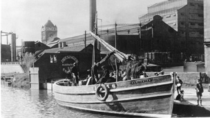 Black and white view of an open decked boat, moored on the Forth and Clyde Canal at the Singer Sewing Machine factory, Clydebank. The large factory building with its tall chimney stacks and the Singer clock tower can be seen in the background.