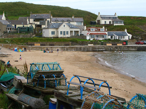 Colour view of narrow beach with the start of a concrete harbour behind and cluster of cottages rises up the hill behind. Lobster pots and related equipment lie piled in the foreground.