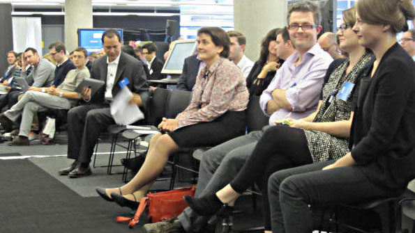 People in suits and serried ranks of chairs, listening to an offscreen speaker.