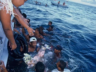 Out in the open sea, men surround a statue of a female, and female hands reach down from a boat towards the offering.