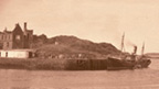 Black and white view of a fishing boat berthed at a simple harbour. The burned out shell of a large house stands in the background.
