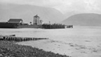 Black and white view from a rocky beach across Loch Linnhe to Ben Nevis. In the middle distance is a quayside featuring a tall, masonry constructed building and a wooden pier.