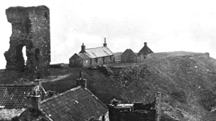 Black and white view of a rocky peninsula on which stand a group of single storey cottages arranged around the ruin of Old Slains Castle.