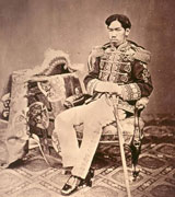 Emperor Meiji, a Japanese man in formal military dress, sits back in a fancy antique chair. A fringed hat like that of a military commander can be seen on the table next to him and the upholstery, carpet and tablecloth are richly patterned