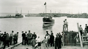 Black and white view of a boat being launched from a slipway into a harbour. A group of men and boys watch from the shore.
