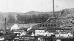 Black and white view of extensive industrial buildings with tall smoking chimney stacks with tall, flat topped hills behind.