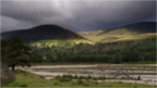 View across winding river on flat valley floor to cloud-darkened hills beyond.