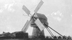 Four sailed timber and stone construction windmill with low stone buildings behind.