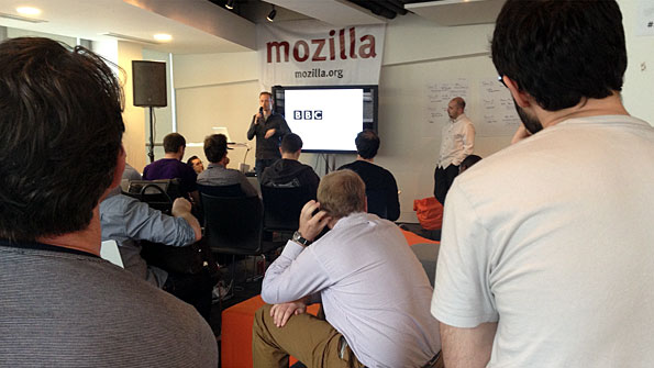 Man speaking in front of a TV displaying the BBC logo, and a Mozilla banner. Other men watch.