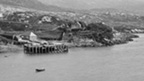 Black and white view across bay to a wooden pier with scattered cottages and other buildings behind.