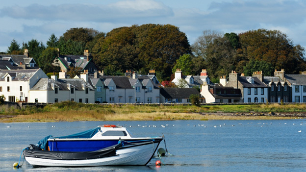A solitary, small boat lies anchored in the bay at Garlieston with the village behind