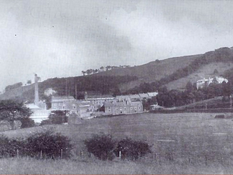 Black and white view of Atheenic Mill buildings and chimney stack, set on a largely grassy slope. There are two short terraces of houses by the mill buildings and a large detached house higher uphill.
