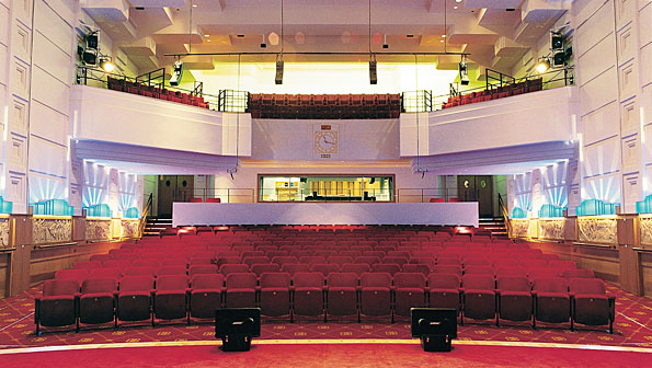 View into a the rows of seating in a theatre. There is a balcony above and a sound booth behind the back row - and art deco lighting around. The carpet is a rich red.