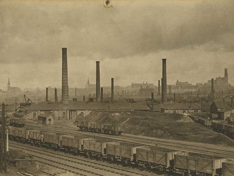 Black and white view of Coatbridge Tinplate Works, dominated by a number of tall, industrial chimney stacks. There is a railway siding in the foreground and the church spires of central Coatbridge can be seen beyond.